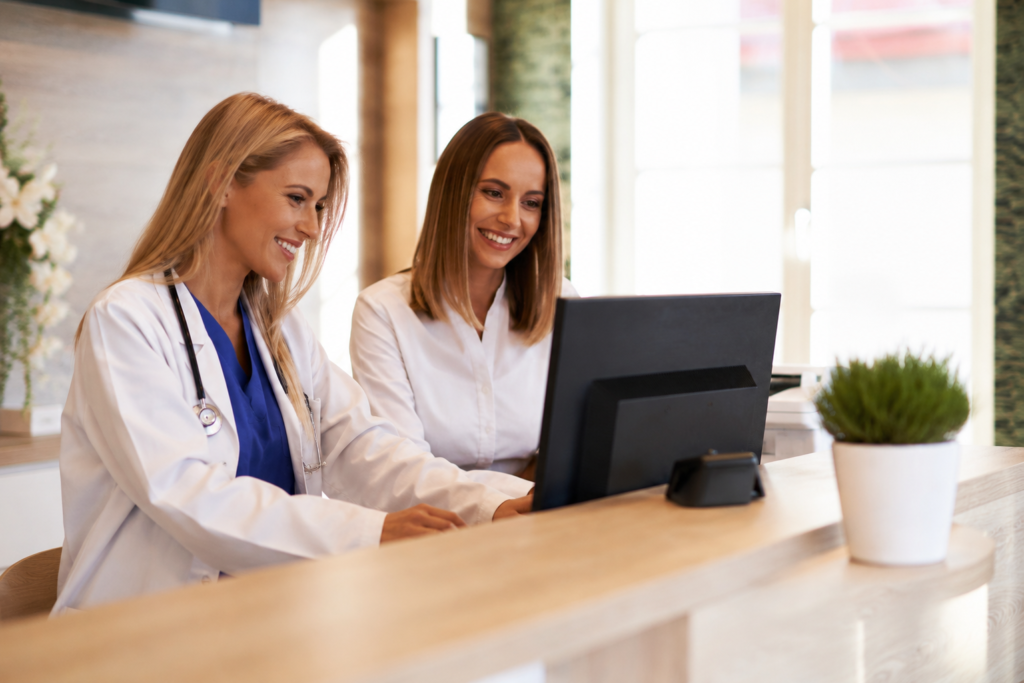 Female doctor stands beside a female administrative assistant while looking at a screen together, smiling.