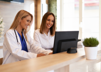 Female doctor stands beside a female administrative assistant while looking at a screen together, smiling. 