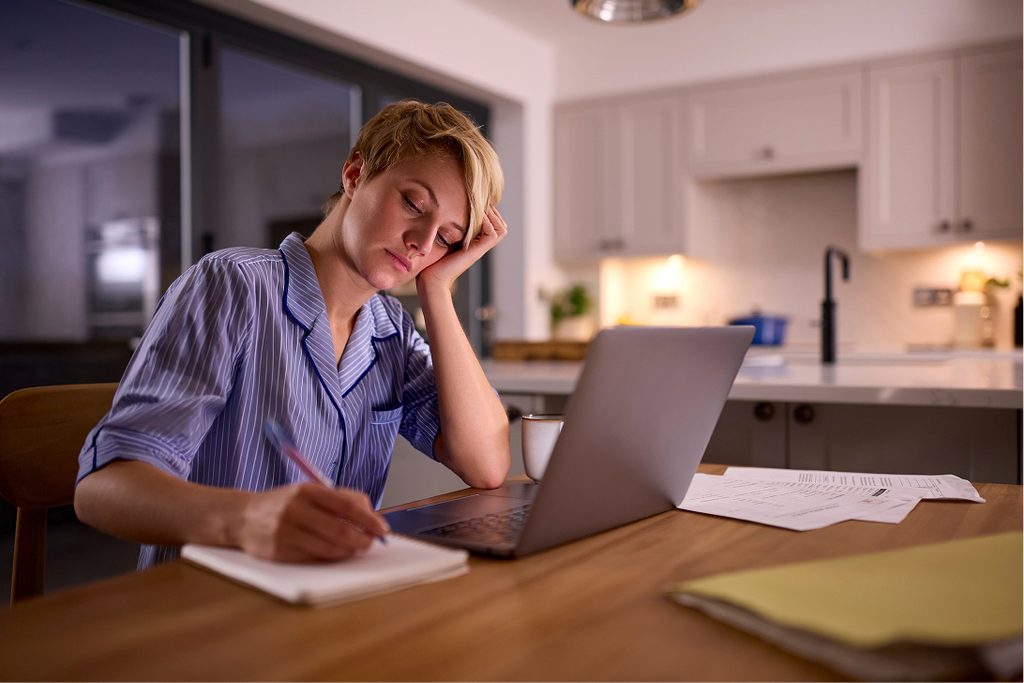 Female doctor sits at a table in her kitchen at night wearing pyjamas, resting her head on her hand as she continues working on her computer into the night.